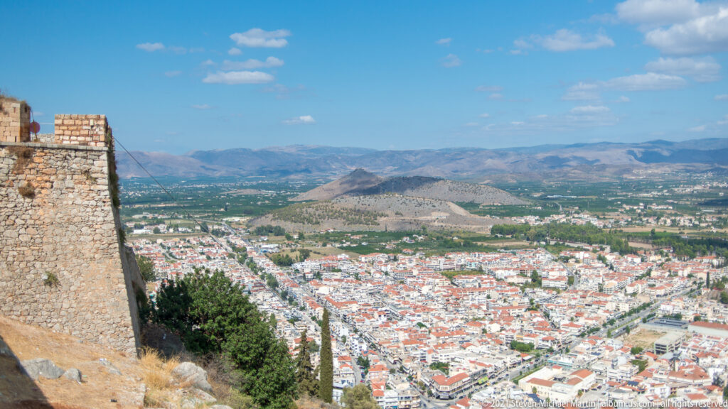 Stone fortress wall beside a high view over dense Nafplio neighborhoods, the Argolid plain, and distant mountains.