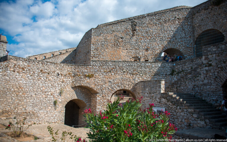 Multi-level Palamidi interior with stone walls, arched openings, stairways, visitors, and flowering shrubs.
