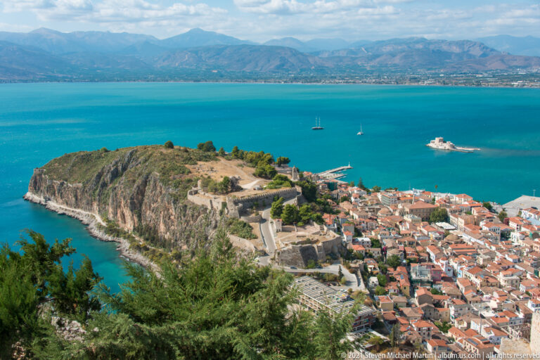 High view over Akronafplia’s rocky fortified promontory, Nafplio’s red roofs, Bourtzi, and the blue Argolic Gulf.