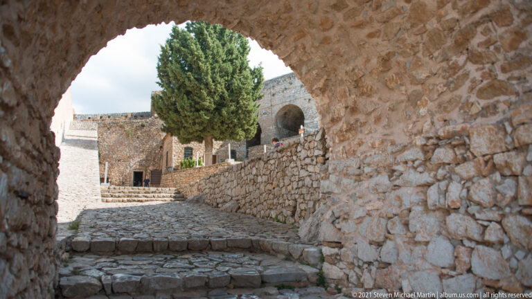 View through a low stone arch toward a cobbled rising passage, cypress tree, terraces, and another arch.