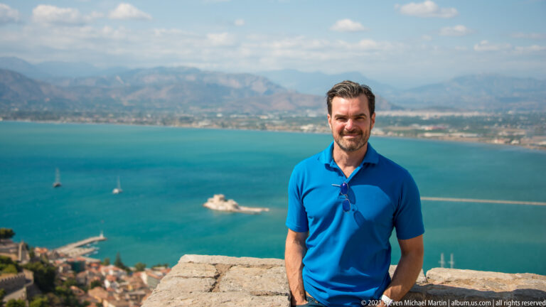 Man in a blue shirt standing at a Palamidi viewpoint with Nafplio harbor and Bourtzi blurred behind him.