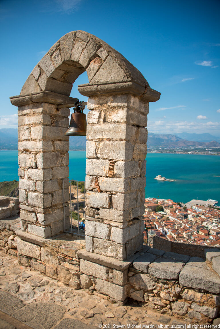 Small arched stone bell frame with a hanging bell above Nafplio and the blue Argolic Gulf.