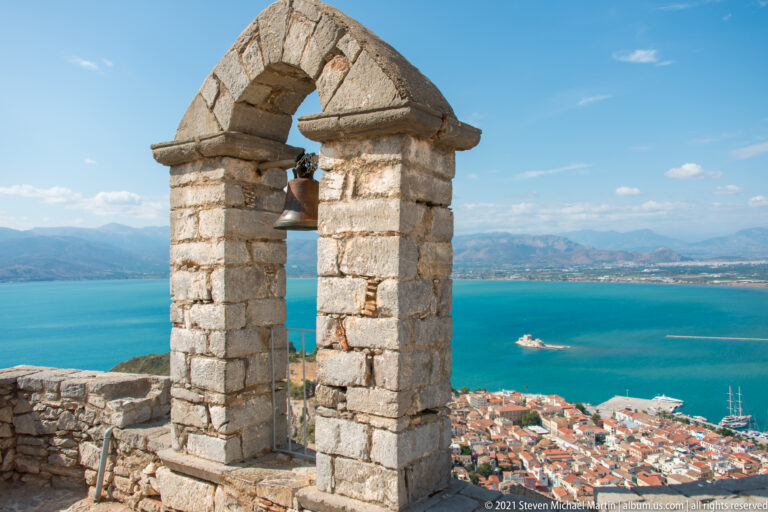 Front view of a stone bell frame at Palamidi, framing Nafplio, the Argolic Gulf, and Bourtzi in the distance.
