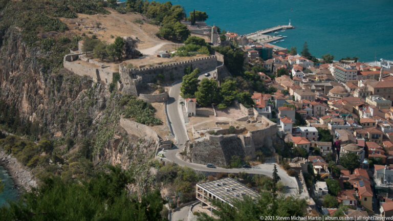 High view of Akronafplia’s stone fortifications, rocky slope, winding road, and the red-roofed edge of Nafplio.