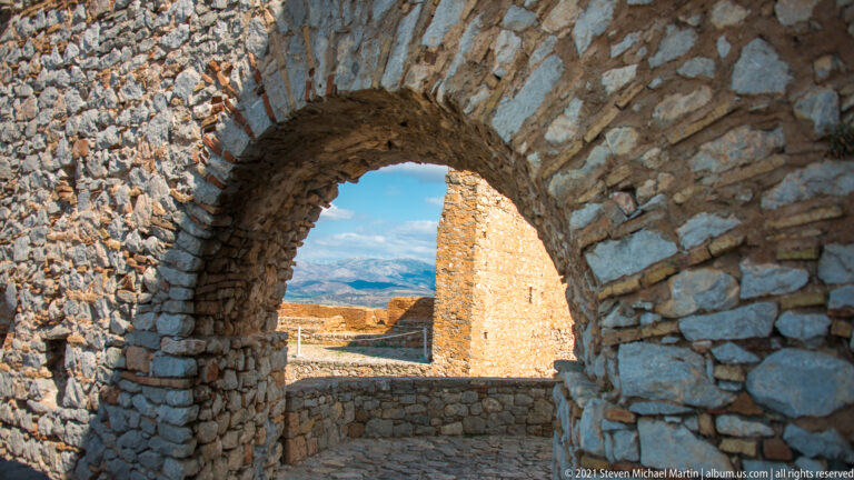 Rough stone arch in shadow framing sunlit fortress walls and distant mountains at Palamidi.