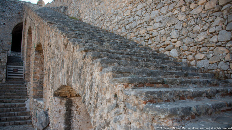 Steep worn stone stairway rising beside a high rough-masonry wall inside Palamidi Fortress.