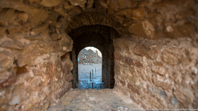 Narrow dark stone corridor with a low vault leading to a small arched doorway and brighter space beyond.