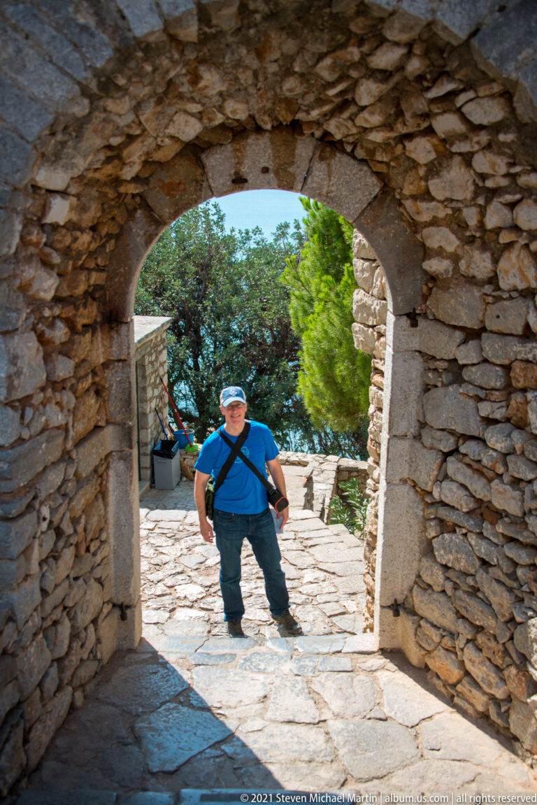 Man standing in a sunlit stone courtyard, framed by a broad shaded arch inside Palamidi Fortress.