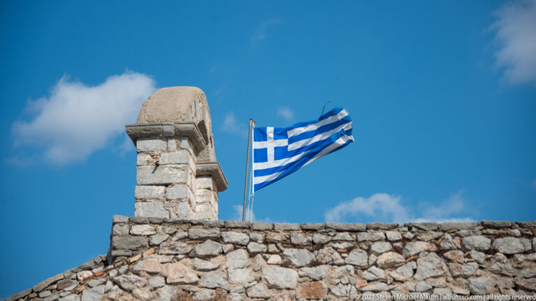 Greek flag flying above a rough stone Palamidi parapet beside a small masonry bell structure.