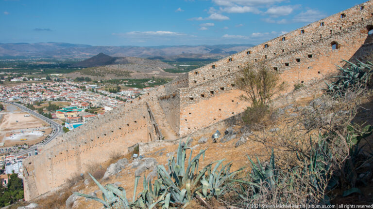Long stone defensive wall descending the Palamidi hillside above Nafplio, with agaves and mountains beyond.