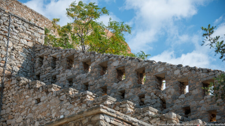 Rough stone Palamidi wall viewed from below, with a row of rectangular defensive openings along its upper edge.