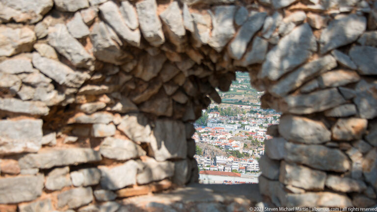 Small opening through thick rough stone masonry framing a distant view of Nafplio below.