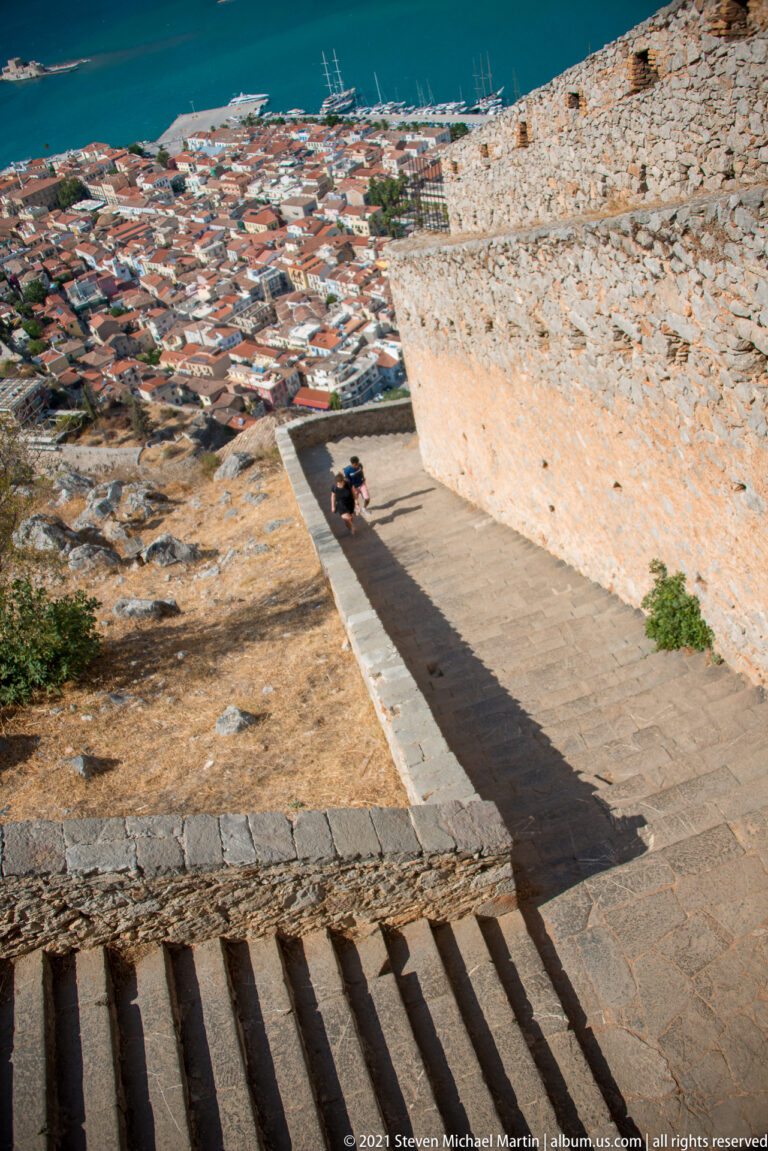 Broad stone steps descending beside a high Palamidi wall toward the red-roofed city and blue water below.