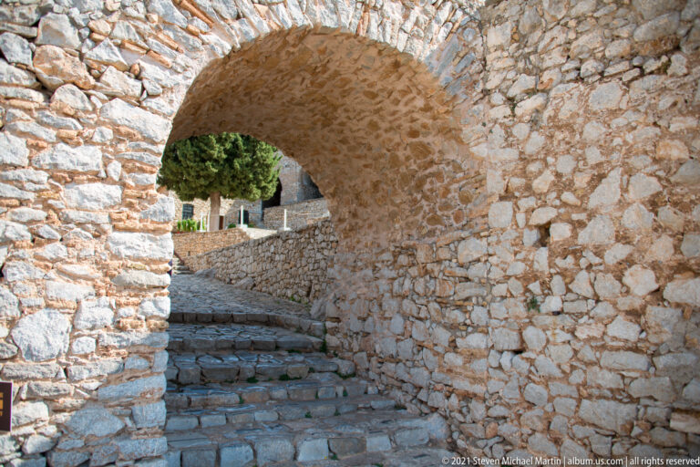 Rough stone barrel vault over cobbled steps leading toward a sunlit exterior area inside Palamidi Fortress.