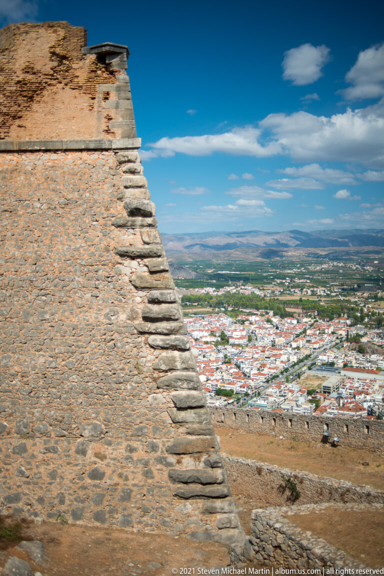 Stepped stone edge of a massive fortress wall with Nafplio and the plain visible far below.