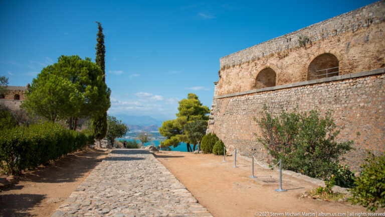 Cobbled path descending beside a high Palamidi wall with large arches, trees and blue water visible ahead.