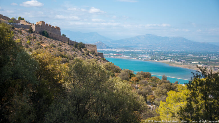 Fortified Palamidi ridge above trees and scrub, overlooking blue coastal water, a broad plain and distant mountains.