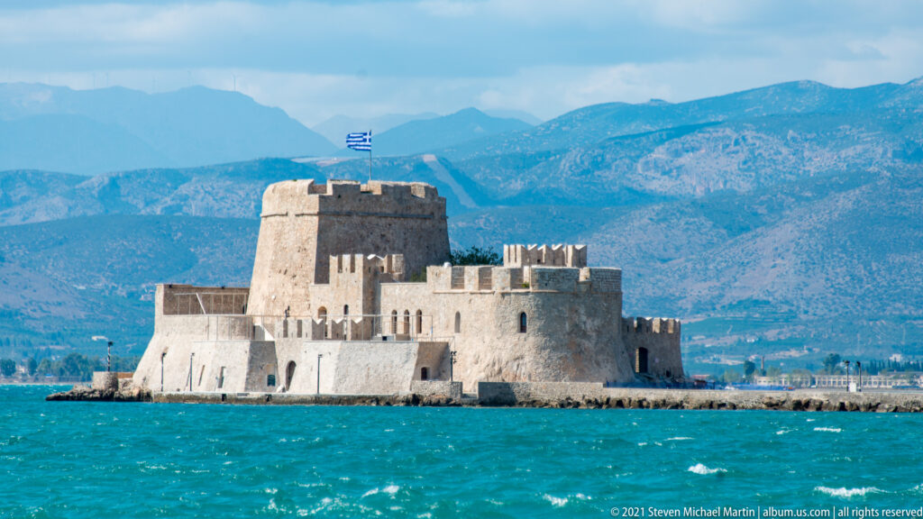 Bourtzi Fortress rising from turquoise water, with crenellated walls, central tower, Greek flag and mountains behind.