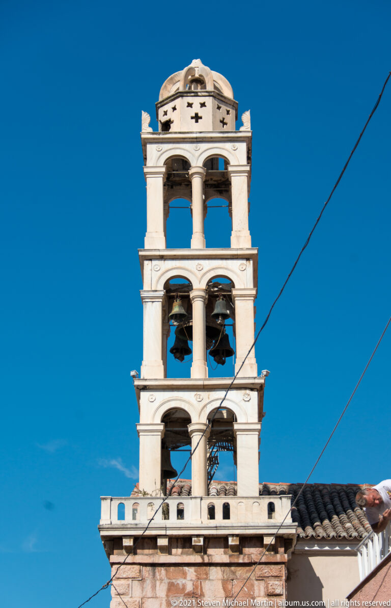 Multi-tiered pale stone bell tower with columns, open arches and several hanging bells against a blue sky.