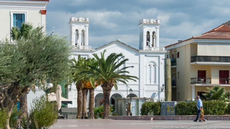 White Orthodox church with twin bell towers behind palms and an olive tree in a Nafplio square.