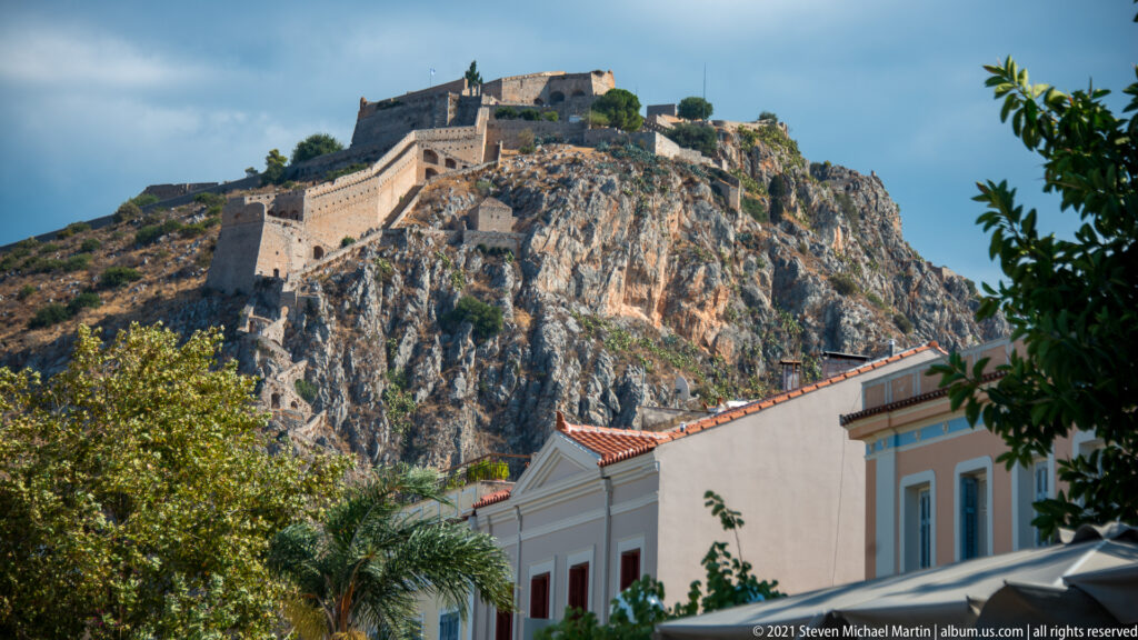 Palamidi Fortress and its walls crowning a steep rocky hill above pastel buildings and trees in Nafplio.