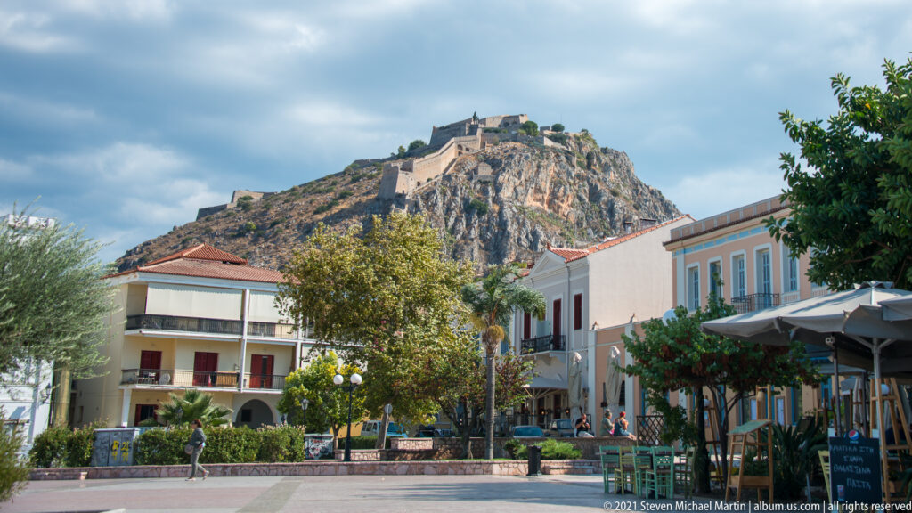 Paved Nafplio square with pastel buildings, café seating and trees beneath the rocky hill crowned by Palamidi Fortress.