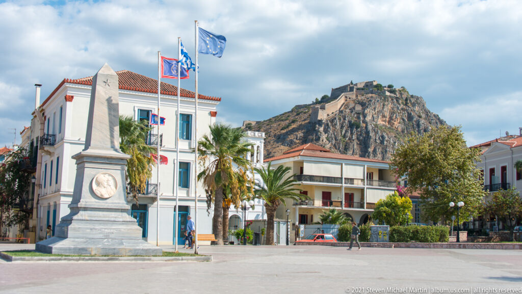 Stone obelisk memorial with a round relief medallion and flags in a Nafplio square, with Palamidi behind.