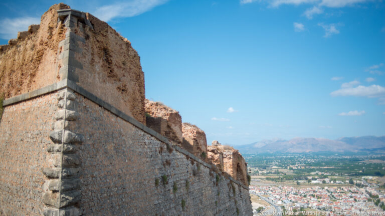 Massive stone fortress wall and projecting upper structure with Nafplio and mountains visible beyond.