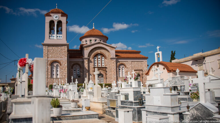 Domed brick-and-stone Orthodox church with bell tower surrounded by white cemetery tombs, crosses and flowers.