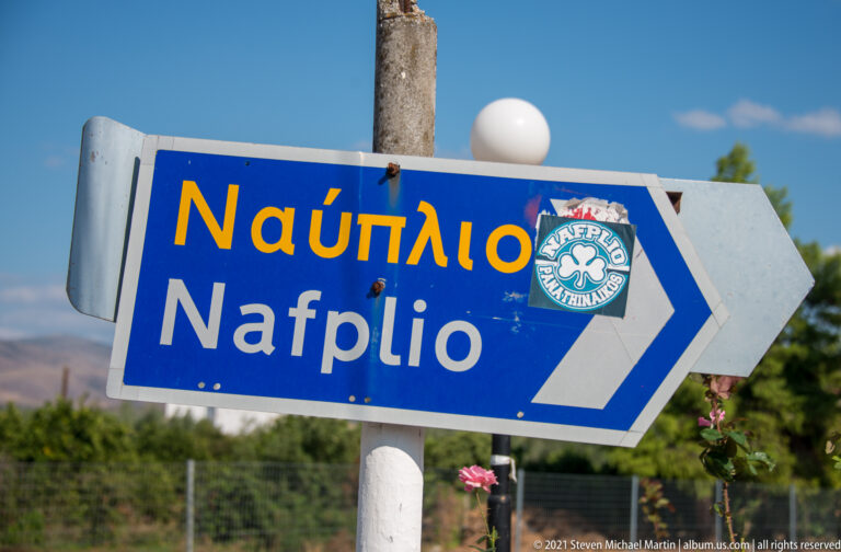 Blue road sign reading “Ναύπλιο / Nafplio” in Greek and Latin script with a white directional arrow.