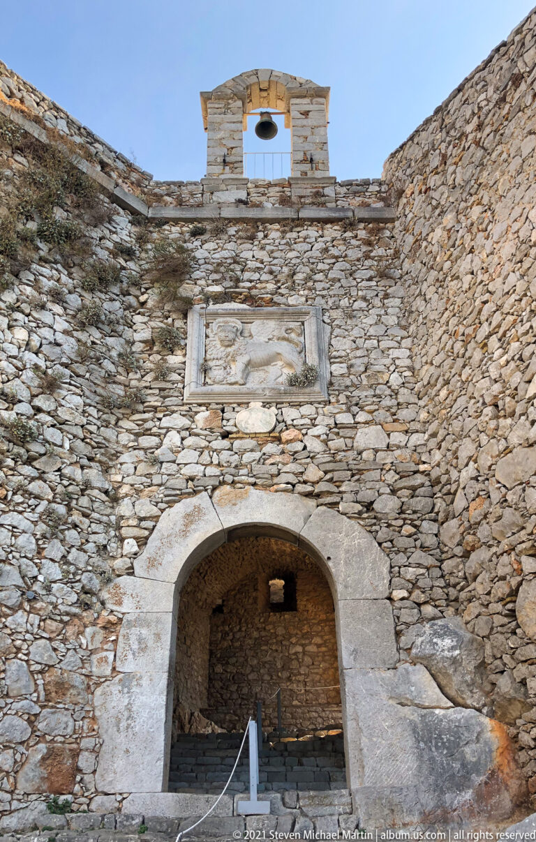 Broad arched opening in a Palamidi stone wall beneath a rectangular relief panel and small bell opening.