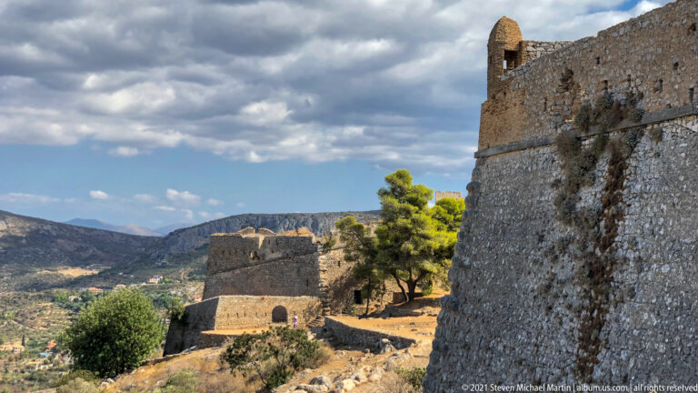 Tall Palamidi wall beside lower bastions and connecting defenses on a dry hillside with pines and mountains.
