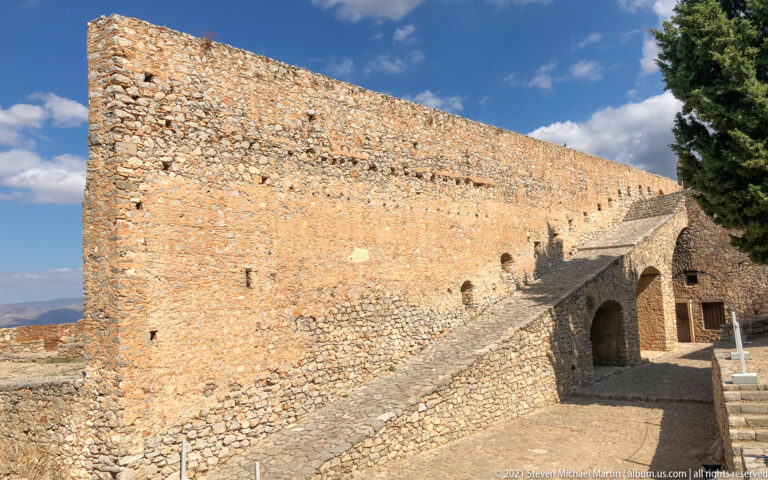 Sloping stone ramp beside a long sunlit Palamidi wall, with arched openings and steps at the far end.