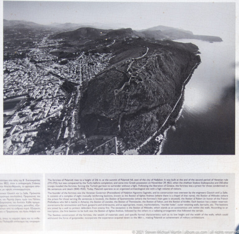 Interpretive panel displaying a black-and-white aerial view of Palamidi’s fortified hill, Nafplio, and the surrounding coast.