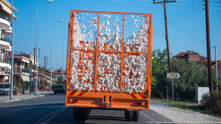Orange mesh-sided road trailer filled with loose white cotton on a street lined with utility poles and low buildings.