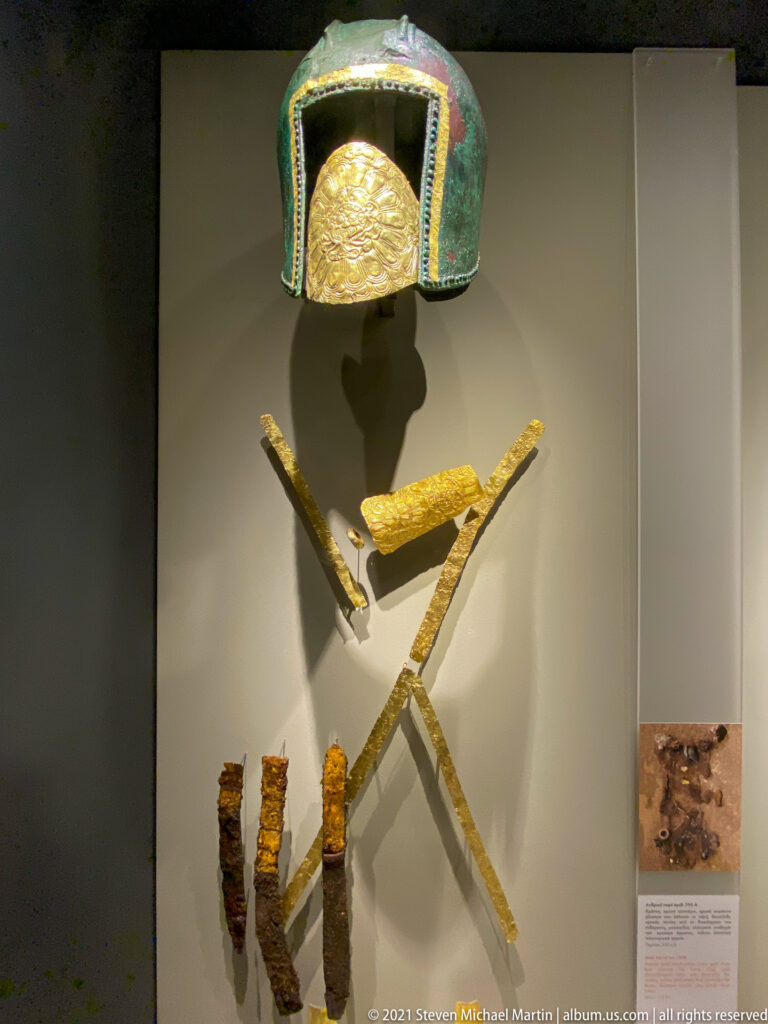Green bronze helmet with gold insert above crossed gold bands and several corroded iron weapon fragments in a museum display.