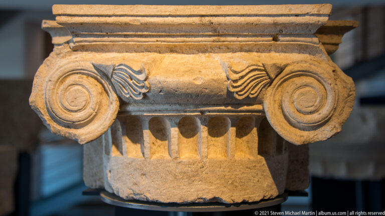 Stone Ionic capital with large spiral volutes, central palmettes, and a row of carved elements beneath the abacus.