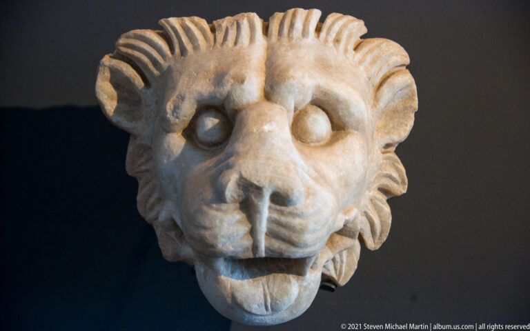 Marble lion head with open mouth, broad muzzle, and deeply carved radiating mane against a dark background.