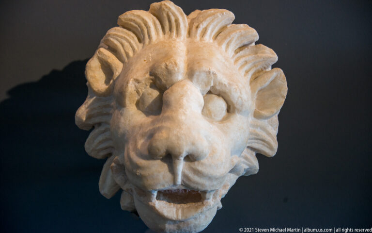Oblique view of a marble lion head with open mouth and layered mane carved around the face.