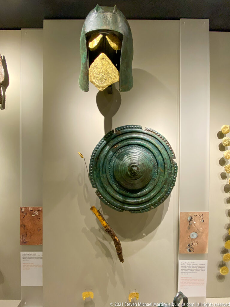 Green-patinated bronze helmet above a large circular shield and smaller gold and metal grave goods.