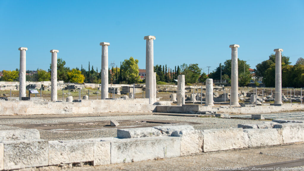 Row of standing stone columns among low excavated walls under a clear blue sky.