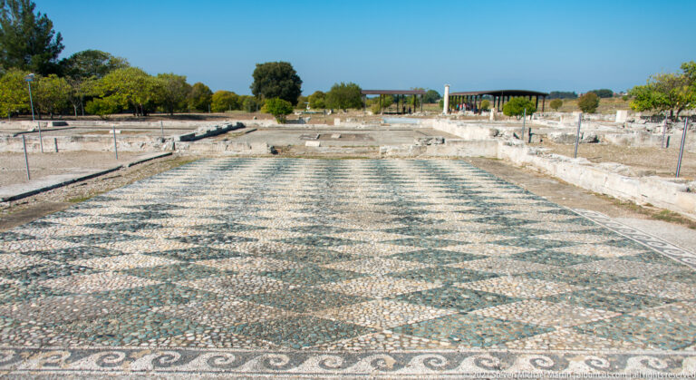 Long in-situ pebble mosaic with repeating geometric pattern and scrolling border amid excavated walls.