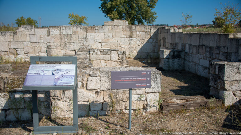Excavated stone-walled area with large masonry blocks, steps, and two visitor interpretation signs.