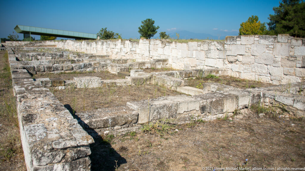 Rectangular excavated building foundations with stepped masonry and a high stone wall along the far side.