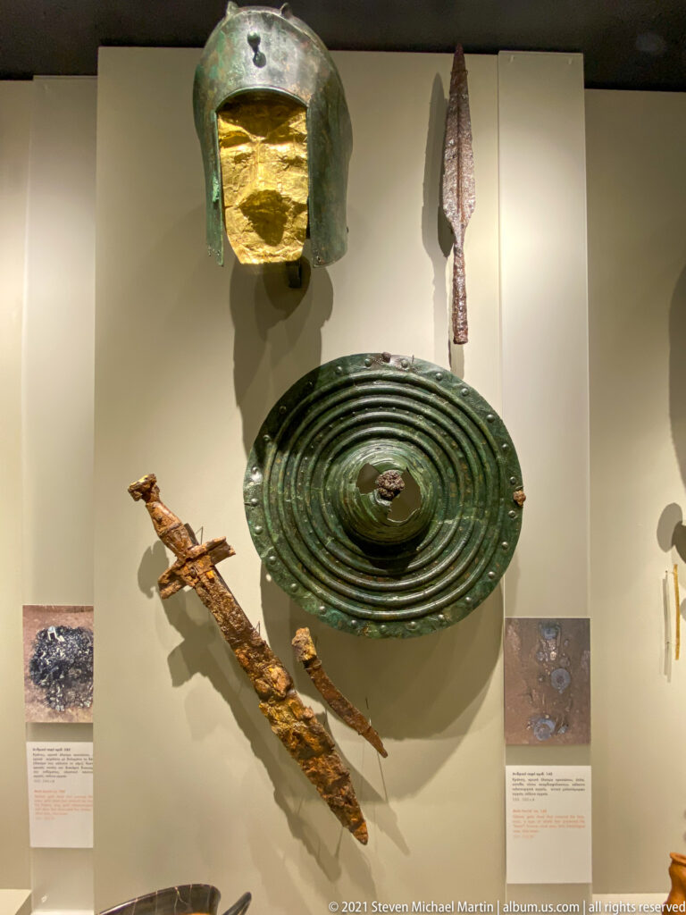 Large ridged bronze shield beneath a bronze helmet, with spearhead and corroded iron weapon fragments arranged around it.