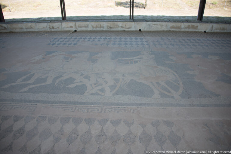 Large in-situ figural pebble mosaic with human and animal forms framed by broad geometric and ornamental borders.