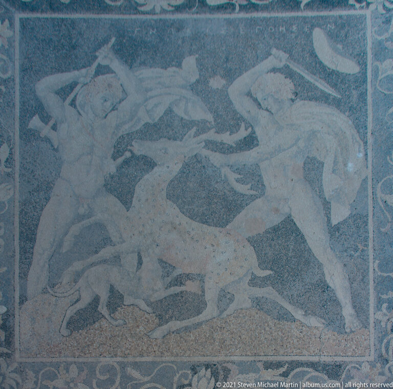 Pebble mosaic of two hunters attacking an antlered stag, with pale figures against a dark ground.