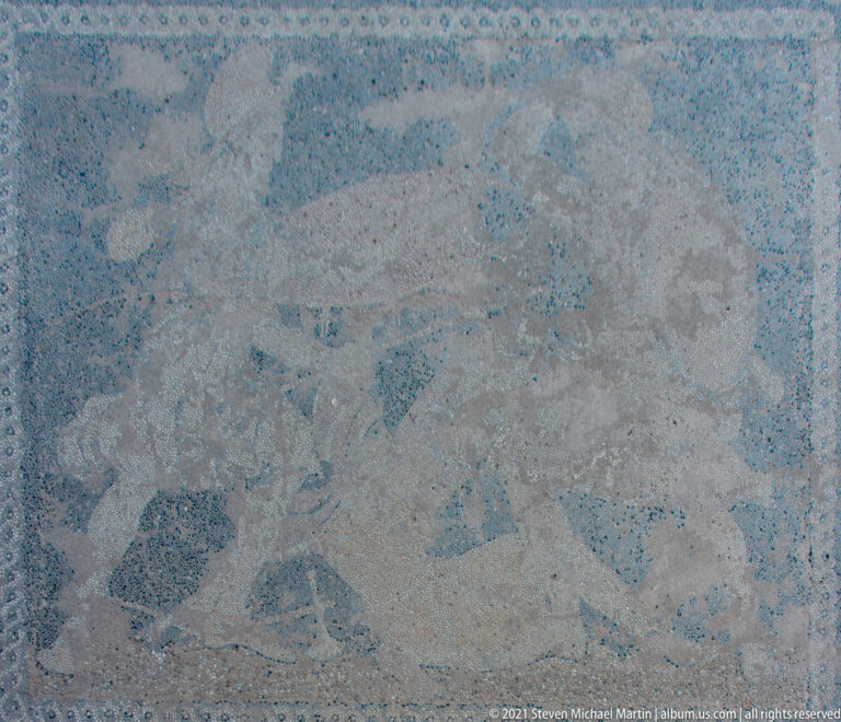 Weathered in-situ pebble mosaic with partially preserved human and animal figures against a dark field.