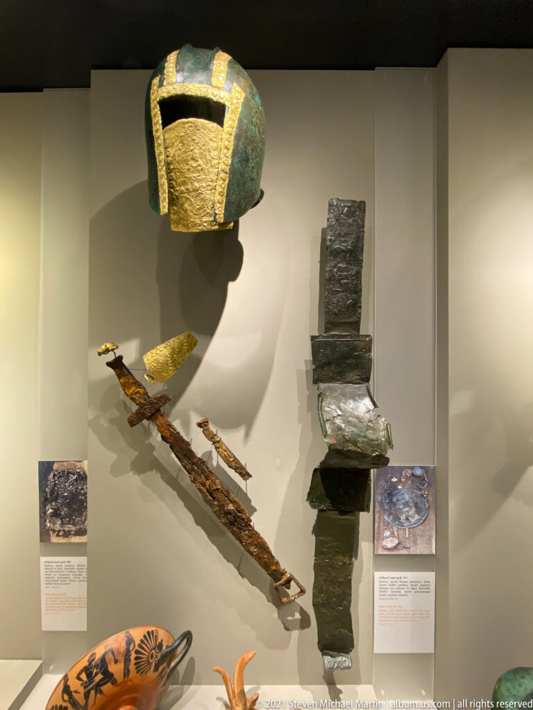 Bronze helmet with gold facing above a decorated corroded blade, beside a broad vertical dark metal fitting in a museum case.