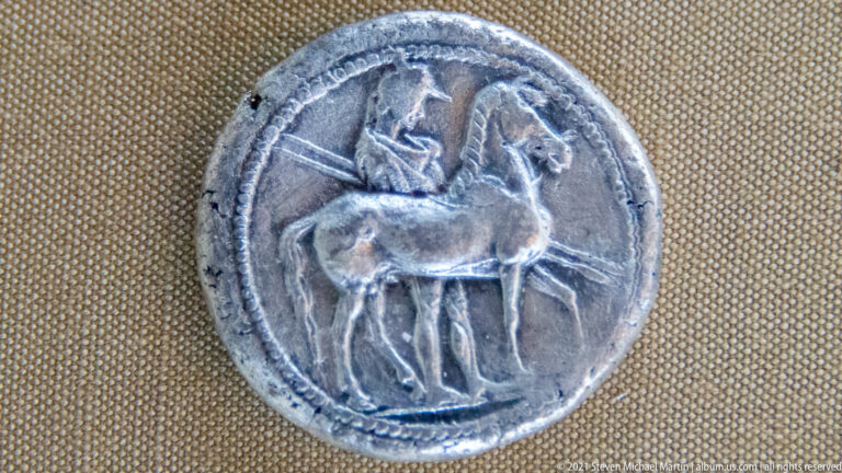 Close-up of a worn silver-toned ancient coin showing a mounted rider on a horse in profile.